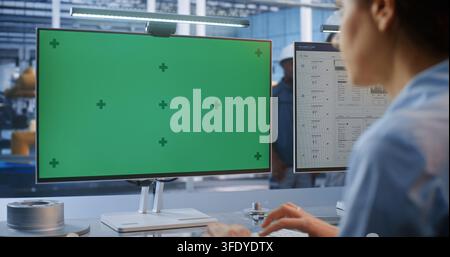 Young Female Technician Sitting at Industrial Workstation Types in Front Green Screen Display with Tracking Markers. Perfect For Compositing Customized Smart Factory Digital Interface. Industry 4.0. Stock Photo