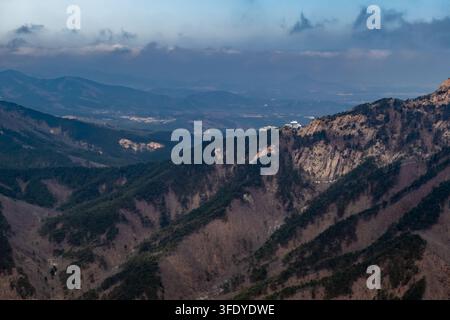 Layered mountain ridges and wooded valleys beneath a cloudy blue sky, with a rocky sunlit peak and distant hills fading into soft evening haze. Stock Photo