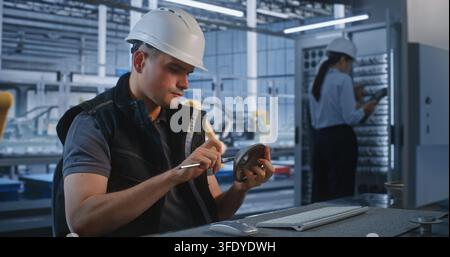 Inside Smart Factory Control Room, Engineer Operates PC while Colleagues Maintain Server Cabinet in Background. Industrial Automation and Server Infrastructure Optimize Manufacturing. Teamwork. Stock Photo