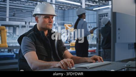 Inside Smart Factory Control Room, Engineer Operates PC while Colleagues Maintain Server Cabinet in Background. Industrial Automation and Server Infrastructure Optimize Manufacturing. Teamwork. Stock Photo