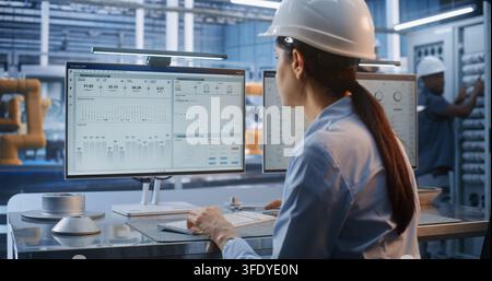 Inside Modern Smart Factory, Female Engineer in Hard Hat Uses Computer to Design 3D Car Chassis. Advanced CAD Software and Automated Robotics Drive Modern Industry 4.0 Manufacturing Efficiency. Stock Photo