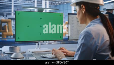 Inside Modern Smart Factory, Young Female Engineer in Hard Hat Operates PC with Chroma Key Green Screen Monitor. Ideal for Compositing Customized Industrial Software and Telemetry. AI Manufacturing. Stock Photo