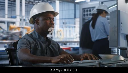 African American Engineer in Hard Hat Types Computer Workstation in Smart Factory Control Room. Background Colleagues Check Server Racks While Robotic Arms Assemble Vehicles. Concept of Robotization. Stock Photo