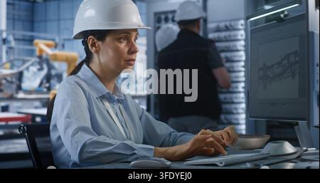 In Modern Smart Plant, Female Professional Engineer in Hardhat Types on PC to Edit 3D Car Chassis Blueprint. Advanced CAD Software and Server Infrastructure Optimize Industry 4.0 Car Production. Stock Photo