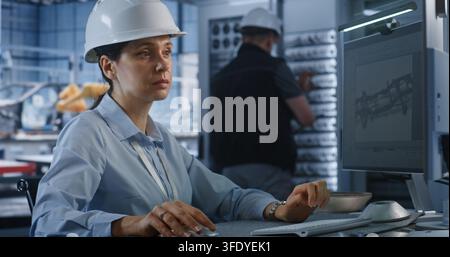 In Modern Smart Plant, Female Professional Engineer in Hardhat Types on PC to Edit 3D Car Chassis Blueprint. Advanced CAD Software and Server Infrastructure Optimize Industry 4.0 Car Production. Stock Photo