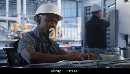 Inside Automated Plant Control Office, African American Specialist Types on PC Keyboard to Manage Production Algorithms. Network Technicians Service Server Hardware in Background. Industry 4.0. Stock Photo