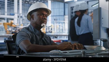 African American Engineer in Hard Hat Types Computer Workstation in Smart Factory Control Room. Background Colleagues Check Server Racks While Robotic Arms Assemble Vehicles. Concept of Robotization. Stock Photo