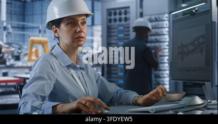 In Modern Smart Plant, Female Professional Engineer in Hardhat Types on PC to Edit 3D Car Chassis Blueprint. Advanced CAD Software and Server Infrastructure Optimize Industry 4.0 Car Production. Stock Photo