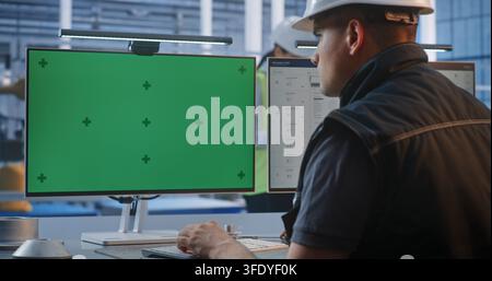 Industrial Worker in Safety Gear Sits at Control Desk Typing, Facing Glowing Green Screen Monitors. Displays Feature Trackers For Clean Chroma Key Compositing in Smart Factory. AI Manufacturing. Stock Photo