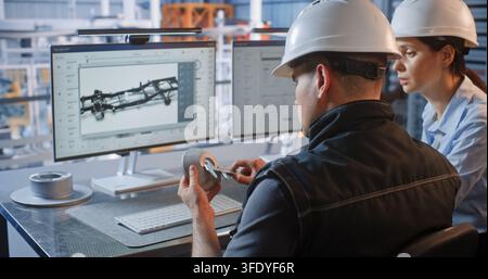 Female Engineer Points at 3D Car Chassis Screen while Discussing Component Design with Colleague Holding Prototype. Teamwork, CAD Software Drive Industry 4.0 Production Efficiency. Team Collaboration. Stock Photo