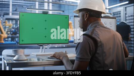 African American Engineer in Hard Hat Sits at Factory Desk Typing in Front of Chroma Key Screen Monitor. Ideal For Compositing Custom Industrial Software User Interfaces. Concept of AI Manufacturing. Stock Photo