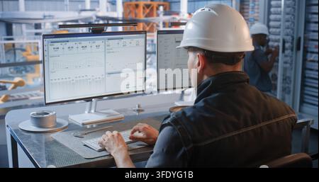 In Glass Control Room, Male Engineer Designs Car Chassis on PC as Robotic Arms Assemble Vehicles on Conveyor Line. Industry 4.0 Technology Drives Factory Efficiency. Concept of Robotization. Stock Photo
