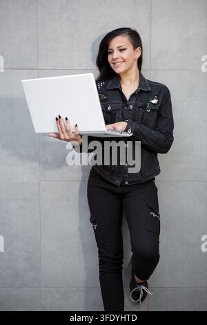 Casual dressed woman standing with laptop and leaning on wall. Stock Photo