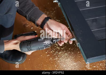 Man securing hardware fitting on wooden door panel with electric drill, close up during installation process. Indoor setting with sawdust and material Stock Photo