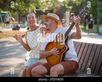 Man playing acoustic guitar while his smiling partner listens beside him in a sunny park Stock Photo