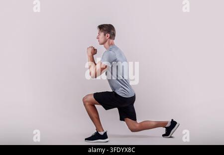 Side view of young fit guy in sportswear doing lunge on light studio background Stock Photo