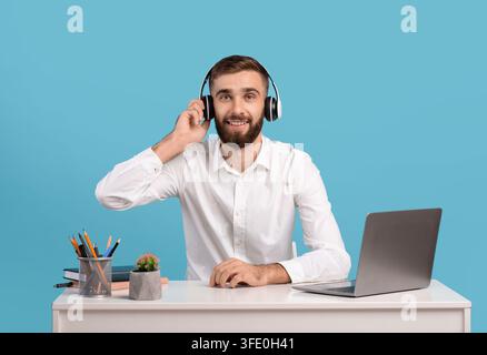 Bearded male helpline operator with headphones and laptop answering customer's call at desk over blue studio background Stock Photo