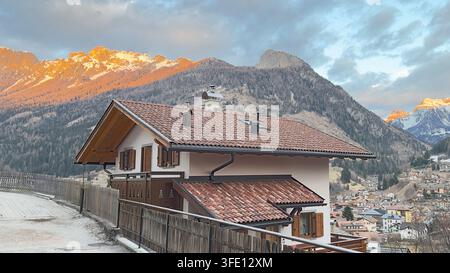 Charming alpine house with tiled roof in a mountain village at sunset with orange glowing dolomites peaks in the background Stock Photo
