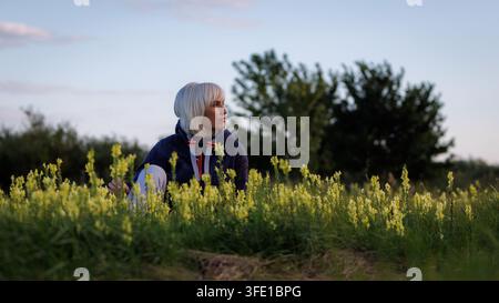A side view portrait of a thoughtful blonde woman in a cozy hoodie sitting in a lush meadow filled with blooming yellow wildflowers, gazing towards th Stock Photo