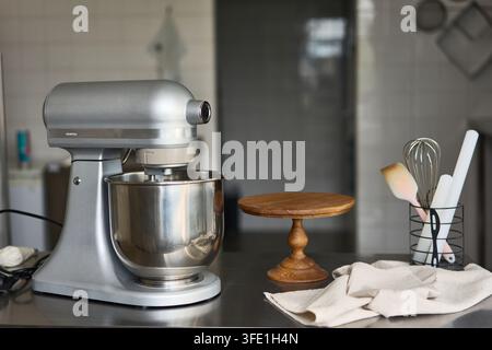 Silver stand mixer with metal bowl on kitchen counter next to wooden cake stand and utensils, with mixer, stand, silver, metal detail, showing Stock Photo