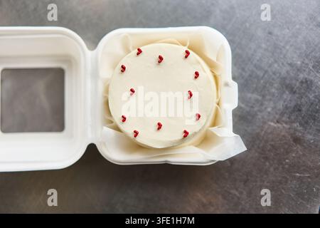 Small round white frosted cake with red piped hearts inside takeaway box on stainless table top, with cake, frosting, white, heart detail, showing red Stock Photo