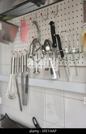 Kitchen pegboard displaying stainless steel utensils knife ladle spoons and whisks hanging on hooks over white tile backsplash organized wall storage Stock Photo