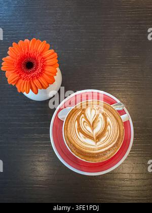 Cappuccino with leaf-shaped latte art in a red ceramic cup beside an orange gerbera flower on a dark wooden table, viewed from above. Stock Photo