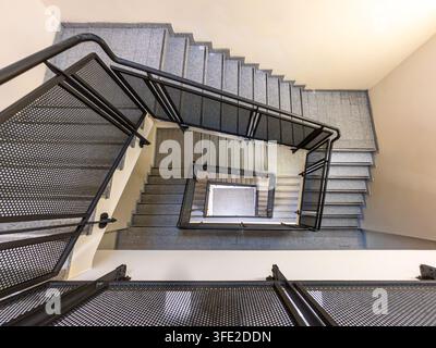 High-angle architectural shot looking down a square-shaped concrete staircase featuring contemporary industrial black mesh handrails Stock Photo