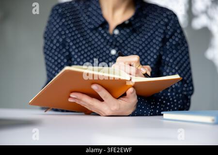 Person's hands holding an open brown leather notebook, writing ideas and plans with a pen, focusing on daily tasks, personal goals, and work strategy on a white desk Stock Photo