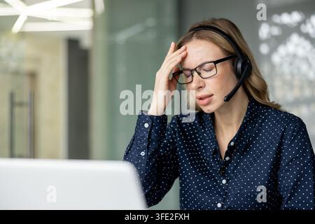 Tired female call center employee wearing a headset and glasses, holding her head, experiencing a severe headache and fatigue from customer support work in the office Stock Photo