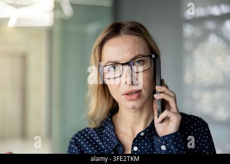 Professional businesswoman wearing eyeglasses having a serious conversation on a mobile phone, discussing work and communicating with clients or colleagues in a contemporary corporate environment Stock Photo