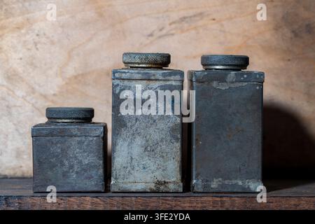 Three vintage rusty metal containers with screw caps on wooden shelf, close up. Row of old aged metallic tins with industrial grunge texture and dark Stock Photo