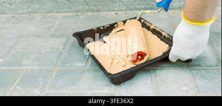 Hand in white glove holds paint roller near tray. Brown paint covers roller and tray surface Stock Photo