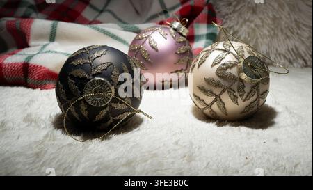 Set of three Christmas tree baubles in black, pink, and cream with sparkling gold glitter fern branch pattern on white plush blanket, soft red plaid t Stock Photo