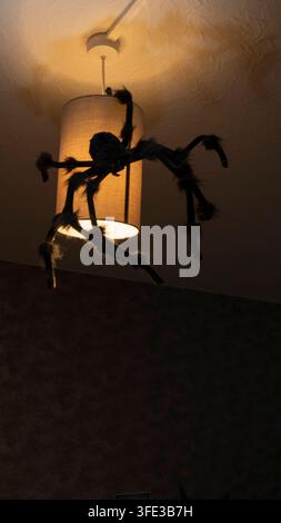 Low angle view of large hairy black spider toy hanging from illuminated ceiling lamp shade with warm glowing light, creepy spooky Halloween home party Stock Photo