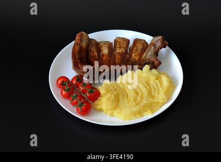 Baked pork ribs with mashed potatoes and cherry tomatoes on a vine in a round plate against a black background. Top view, flat lay. Stock Photo