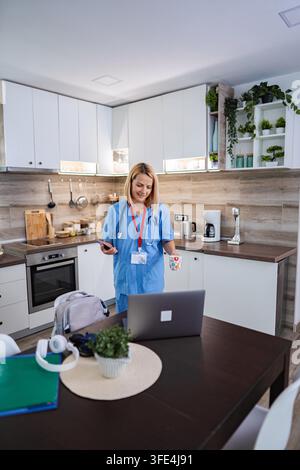 Smiling female doctor in blue uniform drinking coffee and using smartphone and laptop in kitchen Stock Photo