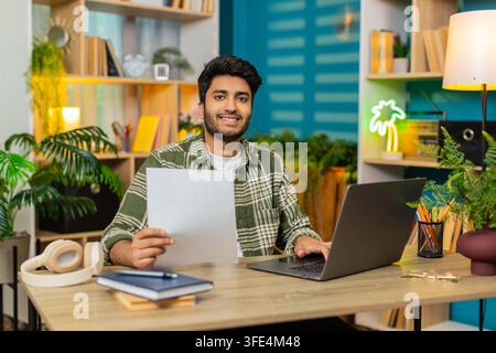 Indian man on home table types on laptop holding important document and smiling at camera in portrait. Arabian guy on home table reviews paperwork for Stock Photo