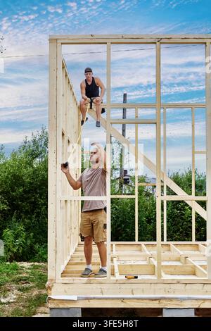 Checking frame dimensions, young builders stretch tape measure between floor framing and top wall beam. Stock Photo