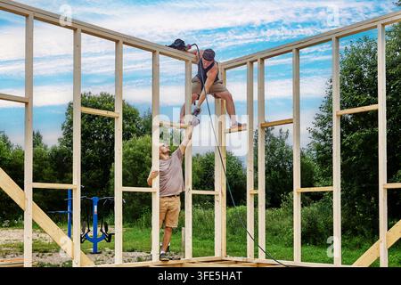 Teamwork in timber construction, with ground worker handing cordless drill to young colleague seated on wall frame. Stock Photo