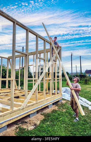 Construction of timber frame house on foundation of concrete blocks, two workers hold timber braces to support wall structure during assembly. Stock Photo