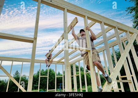 Construction team positions ceiling beams across framework, assembling singlestorey wooden frame house. Stock Photo