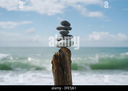 A stack of stones balanced on top of a weathered log against a blurry ocean and sky background. Stock Photo