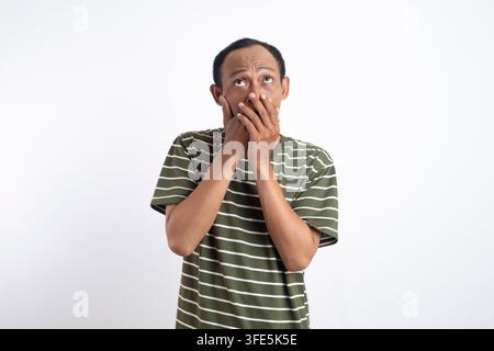 Thoughtful man wearing a green shirt with white stripes looking upwards with a pensive expression, hand on chin, isolated on plain light gray backgrou Stock Photo