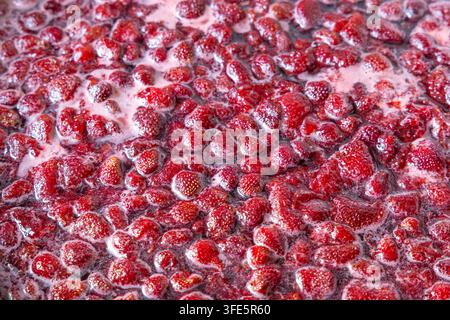 Extreme close-up captures whole strawberries bubbling in thick, glossy red syrup. The scene highlights the cooking process for homemade jam or preserv Stock Photo