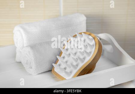 Wooden and silicone scalp massager and rolled towels on a white wooden tray in a home bathroom. Stock Photo