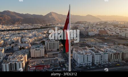 Aerial view of the national flag of Oman flying on a tall flagpole over the urban landscape of Muscat with mountains in the background during golden h Stock Photo