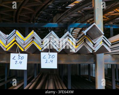 Close-up view of stacks of gray L-shaped metal beams with yellow stripes on industrial shelving. Two white tags with handwritten text hang from the be Stock Photo