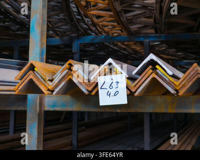 Close-up view of stacked L-shaped metal beams on blue shelving unit in industrial setting. Beams show rust and have identification tag attached, with more metal stock visible in background. Stock Photo