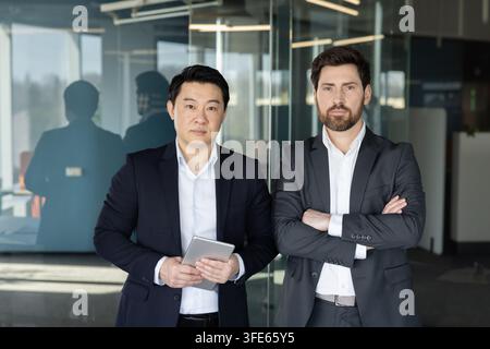 Two diverse businessmen standing confidently in a contemporary office setting, one holding a digital tablet, representing partnership, innovation, and Stock Photo